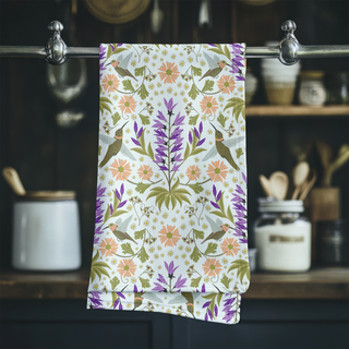 Tea towel with illustration of flowers and hummingbirds hanging on a rack. Sold by JeanC Studio