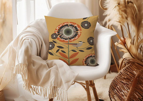 image of a throw pillow on a chair. the pillow has a gold background and one large stylized orange and tan flower. There is also a tan and orange bird sitting on a stem. Smaller brown and dark brown flowers surround it. 