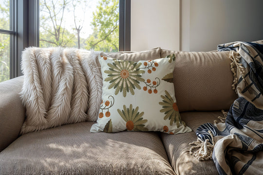 closeup image of a throw pillow on a sofa. the pillow has several flowers in a light warm green, dark green and white with stylized petals and orange berries. 