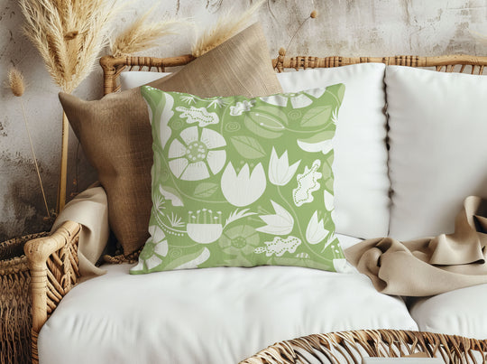 closeup image of a throw pillow on a rattan sofa. the pillow has a lot of abstract flowers in a palette of cream  and pale green. There are abstract green and white leaves surrounding it. There are also yellow green leaves in the background. The background is a fresh medium green color. It's a very summery or spring pillow cover.